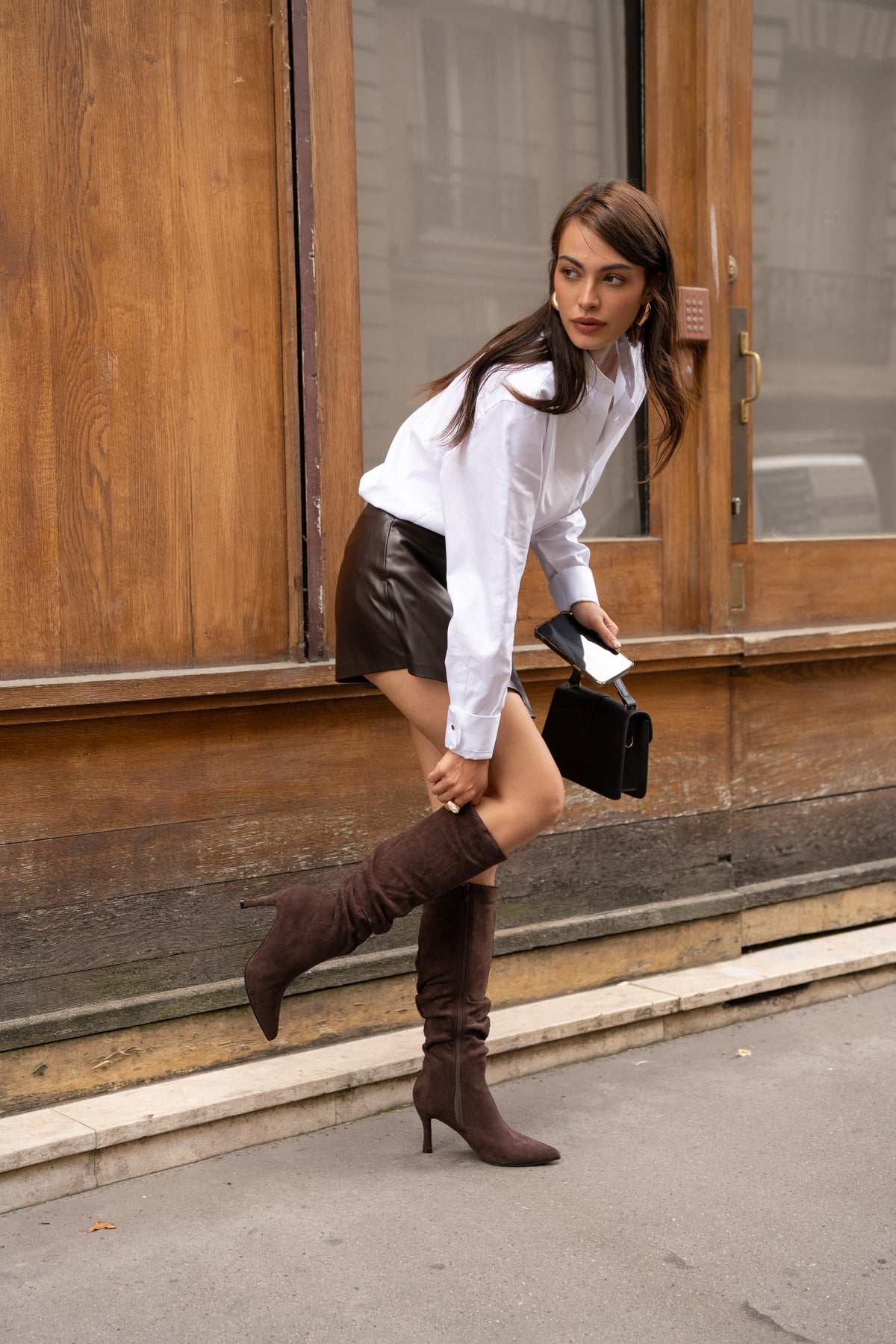 Une femme en chemise blanche et jupe noire ajuste une botte marron à talon devant une vitrine.